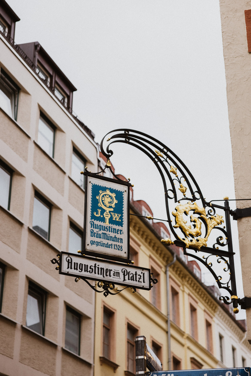 Auf diesem Foto sieht man ein Wappen, das an der Ecke der Außenfassade des Lokals befestigt wurde und herausragt. An goldenen und schwarzen Stahlträgern hängt das Wappen von Augustiner Bräu München. Auf dem blau-goldenen Wappen ist folgender Schriftzug zu lesen: Augustiner Bräu München gegründet 1328. Darunter ist noch ein Schild befestigt, auf dem steht „Augustiner am Platzl“. Im Hintergrund sind ein grauer Himmel und eine Häuserfassade mit Fenstern zu erkennen. Das Bild wurde von unten aufgenommen.