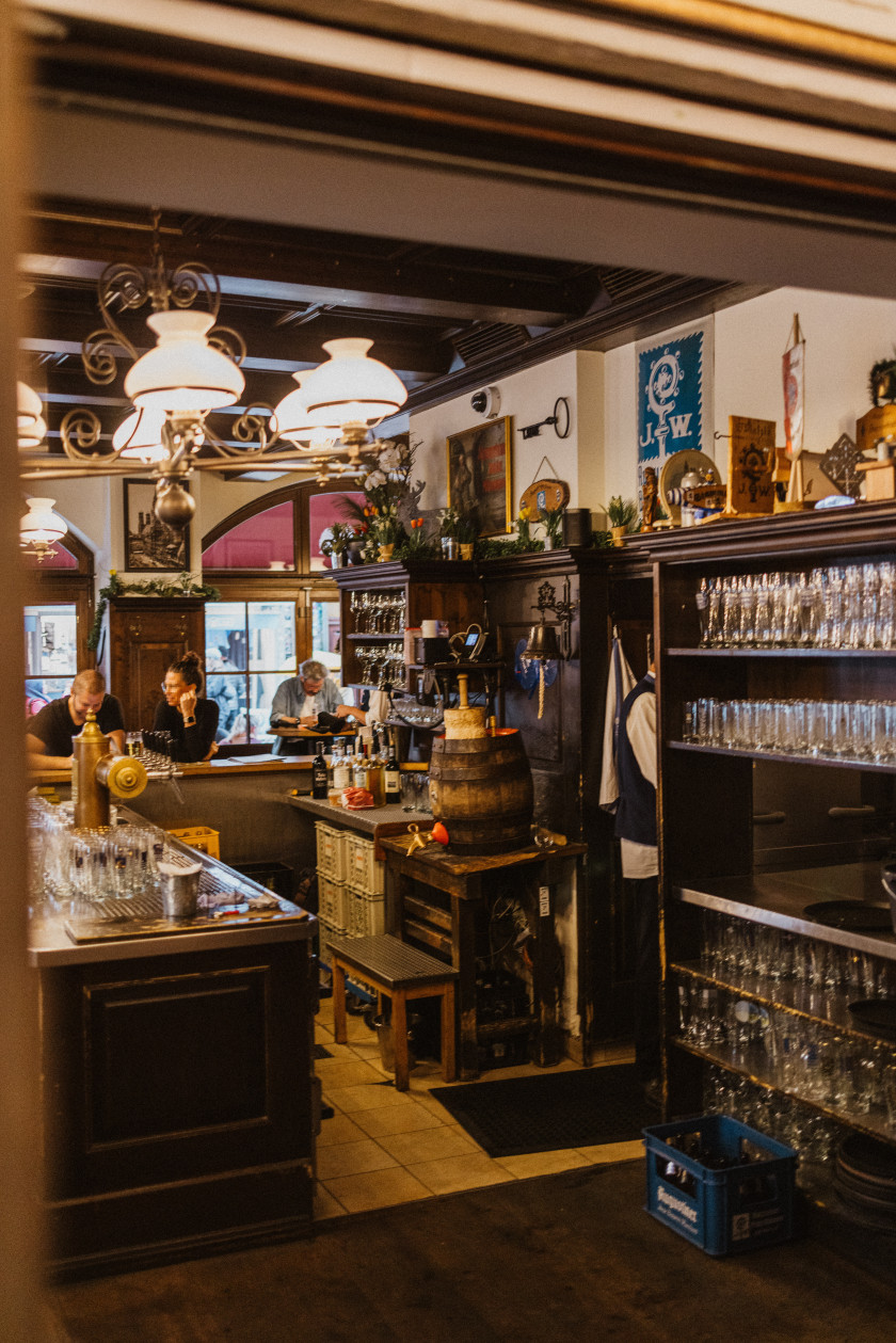 Auf diesem Foto sieht man einen Blick in das Wirtshaus und hinter die Theke. Rechts im Bild ist ein Regal mit leeren Gläsern. Auf dem Regal stehen verschiedene Dekorationen. Links im Bild ist seitlich die Arbeitsfläche hinter dem Tresen mit einer Zapfanlage und leeren Gläsern zu sehen. Neben dem Regal auf der rechten Seite ist von hinten ein Mann zu erahnen, der in der Tür lehnt. Daneben sind ein Holzfass sowie weitere Utensilien für die Bar. Von der Decke hängen Lampen. Der Boden ist zum Teil aus dunklem Holz und der Teil hinter der Bar ist mit braunen Fliesen versehen. Im Hintergrund sieht man zwei Personen zusammen an der Bar sitzen. Noch weiter im Hintergrund rechts sitzt ein Mann vor den Fenstern des Lokals.