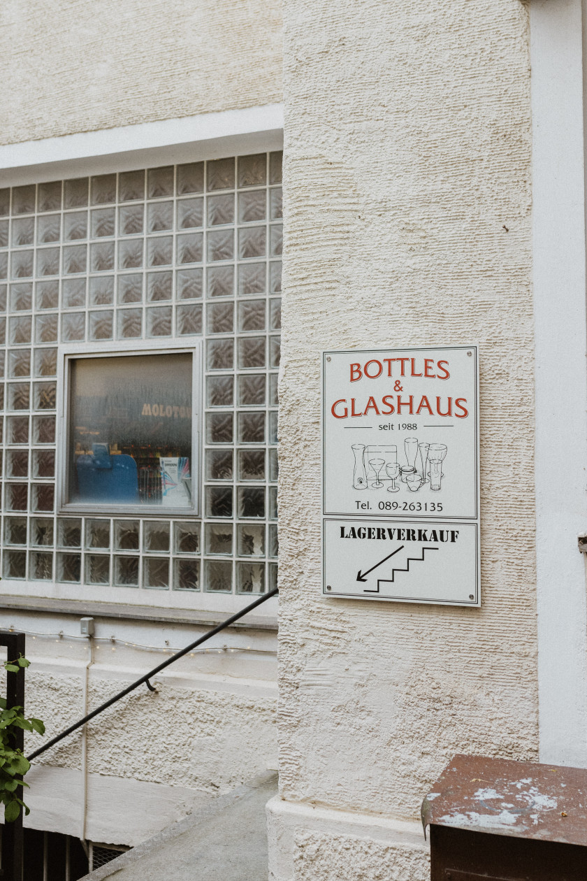 Auf diesem Bild sieht man den Beginn vom Treppenabgang zu dem Geschäft. Dort ist ein Schild „Bottles & Glashaus, seit 1988, Tel. 089-263135“ und darunter ein Schild „Lagerverkauf“ mit einem Pfeil, der nach unten zeigt. Außerdem sieht man das Treppengeländer und das Fenster des angrenzenden Gebäudes.