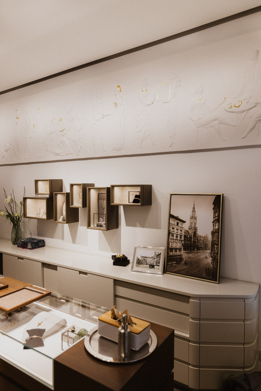 Auf diesem Foto sieht man einen Blick in das Geschäft. Oben an der Wand ist ein Stuck-Wandbild in Weiß mit goldenen Details zu erkennen. Darunter sieht man mehrere Vitrinen an der Wand hängen. Darunter steht ein weißes Sideboard an der Wand. Auf dem Sideboard stehen Bilder, eine Vase und weitere Gegenstände, die schwer zu erkennen sind. Vor dem Sideboard im Raum steht ein Verkaufstresen, der zum Teil aus Glas ist. In dem Tresen sind Schmuckstücke durch das Glas zu erkennen. Auf dem Verkaufstresen steht außerdem ein Tablett mit einer Box mit Tüchern und einem Seifenspender.