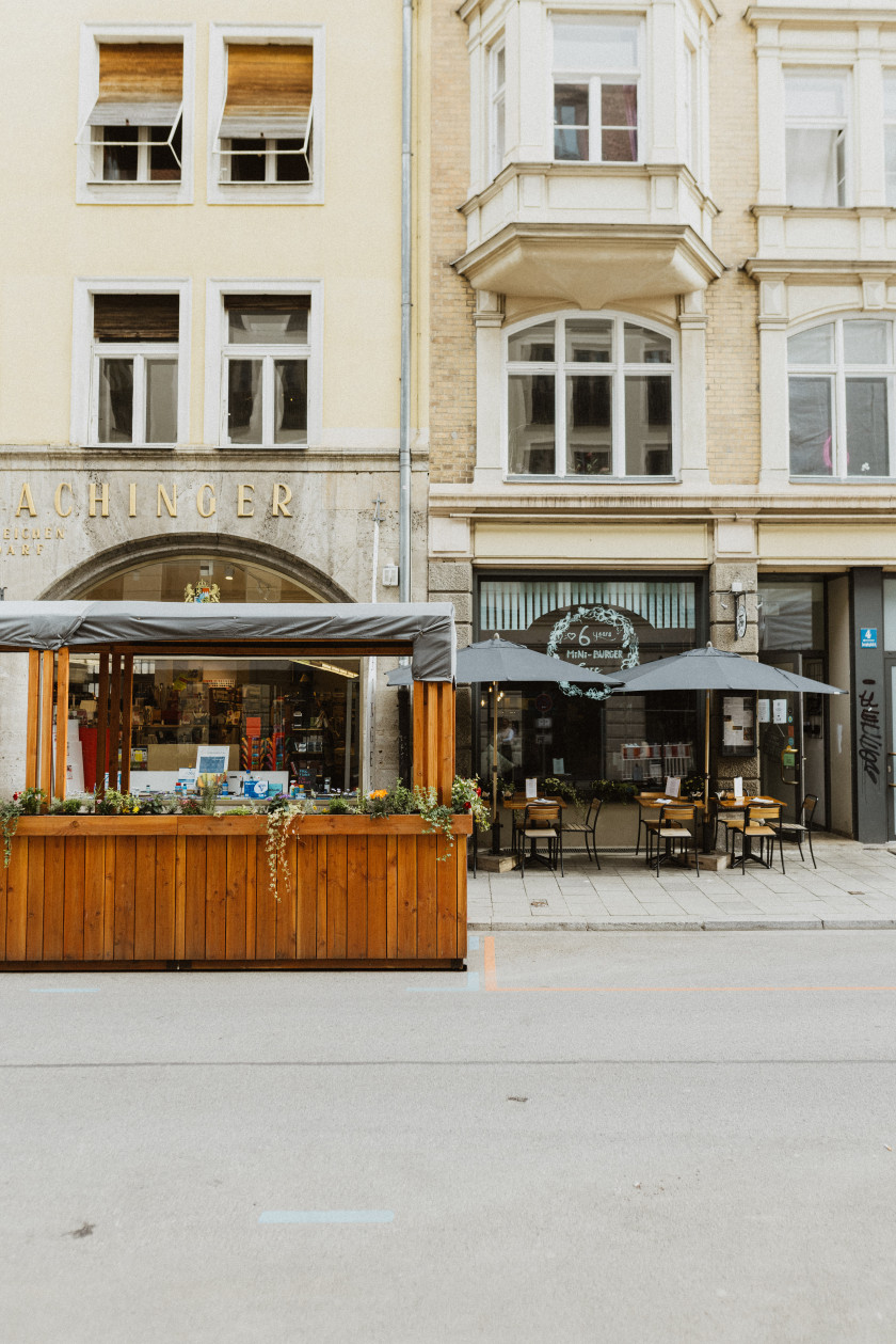Auf diesem Foto sieht man das Restaurant von Außen. Das Foto wurde von der Straße vor dem Laden aufgenommen. Links im Bild ist vor dem Restaurant auf der Straße vor dem Gehweg ein Schanigarten zu sehen. Dieser ist aus Holz und außen sind Blumenkästen angebracht. Darüber ist eine Plane gespannt. Hinter dem Schanigarten ist ein Gebäude, auf dem der Schriftzug „ACHINGER“ zu lesen ist. Das Gebäude ist gelb. Im rechten Teil des Bildes sieht man das Restaurant. Davor sind Stühle und Tische mit Sonnenschirmen auf dem Gehweg aufgebaut. Rechts daneben im Foto ist die Eingangstüre. Im Vordergrund des Fotos ist noch ein Teil der Straße zu erkennen.