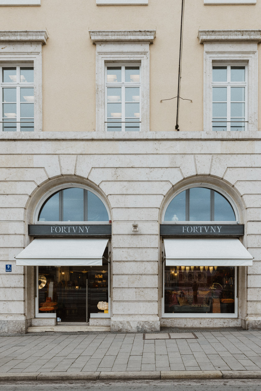 Auf diesem Foto sieht man die Außenfassade von Fortuny München aus der Perspektive der Briennerstraße. Das Geschäft befindet sich in einem älteren Haus und die Außenfassade wirkt edel. Links und rechts sind jeweils ein Schaufenster mit Überdachungen und eines mit Eingang. Über der Überdachung steht der Schriftzug von Fortuny.