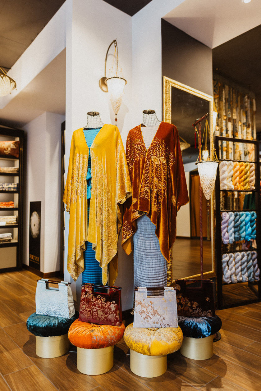 Im Fokus hängen hier zwei Samt-Abendmäntel an Schaufensterpuppen. Der eine ist gelb, der andere rot. Beide sind sehr elegant und gemustert. Davor stehen drei Taschen auf jeweils einem Hocker und werden präsentiert. Angestrahlt wird das Ganze von einer Seidenleuchte. Im Hintergrund sind ein Spiegel, eine Lampe und Wollstränge zu sehen.