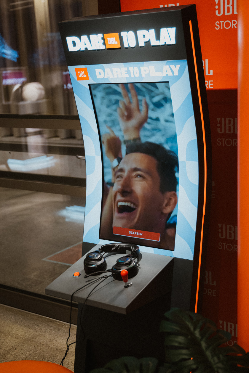 Auf diesem Foto soll eine Spiele-Konsole dargestellt werden. Auf dem Pult liegen Kopfhörer und es sind ein roter Joystick und ein Knopf zu sehen. Hinter dem Pult ist ein Bildschirm zu sehen, auf dem ein Mann mit jubelndem Gesicht und der Schriftzug „JBL DARE TO PLAY“ gezeigt wird. Dieser Schriftzug ist auch über dem Bildschirm zu lesen. Im Hintergrund sind ein roter Aufsteller und eine Scheibe zu sehen.