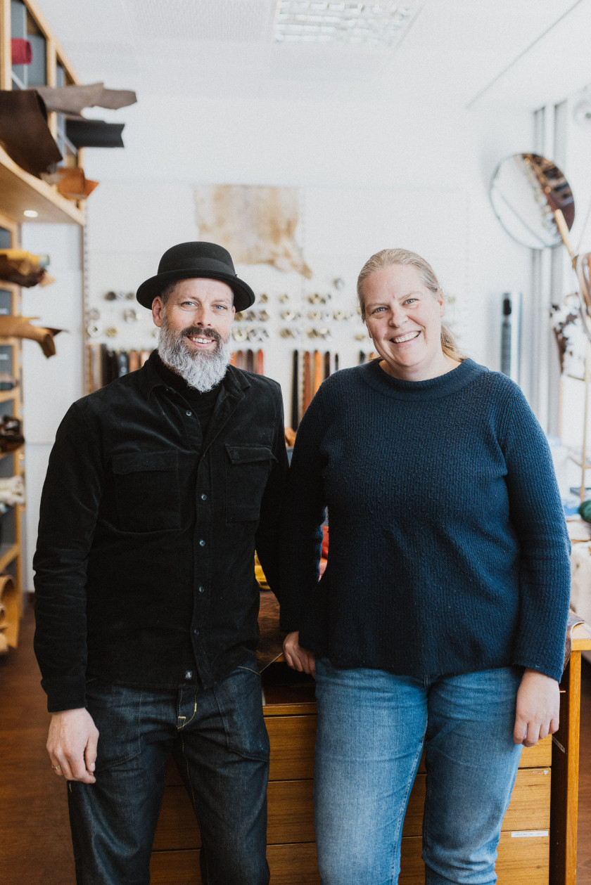 Auf dem Bild sieht man die beiden Inhaber. Links auf dem Bild ist Tobias Müller zu sehen und rechts im Bild ist seine Frau Ines Müller zu sehen. Tobias Müller ist in Schwarz gekleidet und trägt einen schwarzen Hut. Ines Müller trägt blaue Jeans und einen blauen Pullover. Die beiden lächeln in die Kamera und stehen im Laden. Im Hintergrund sieht man das Geschäft und verschwommen Gürtel, Schnallen und Leder.