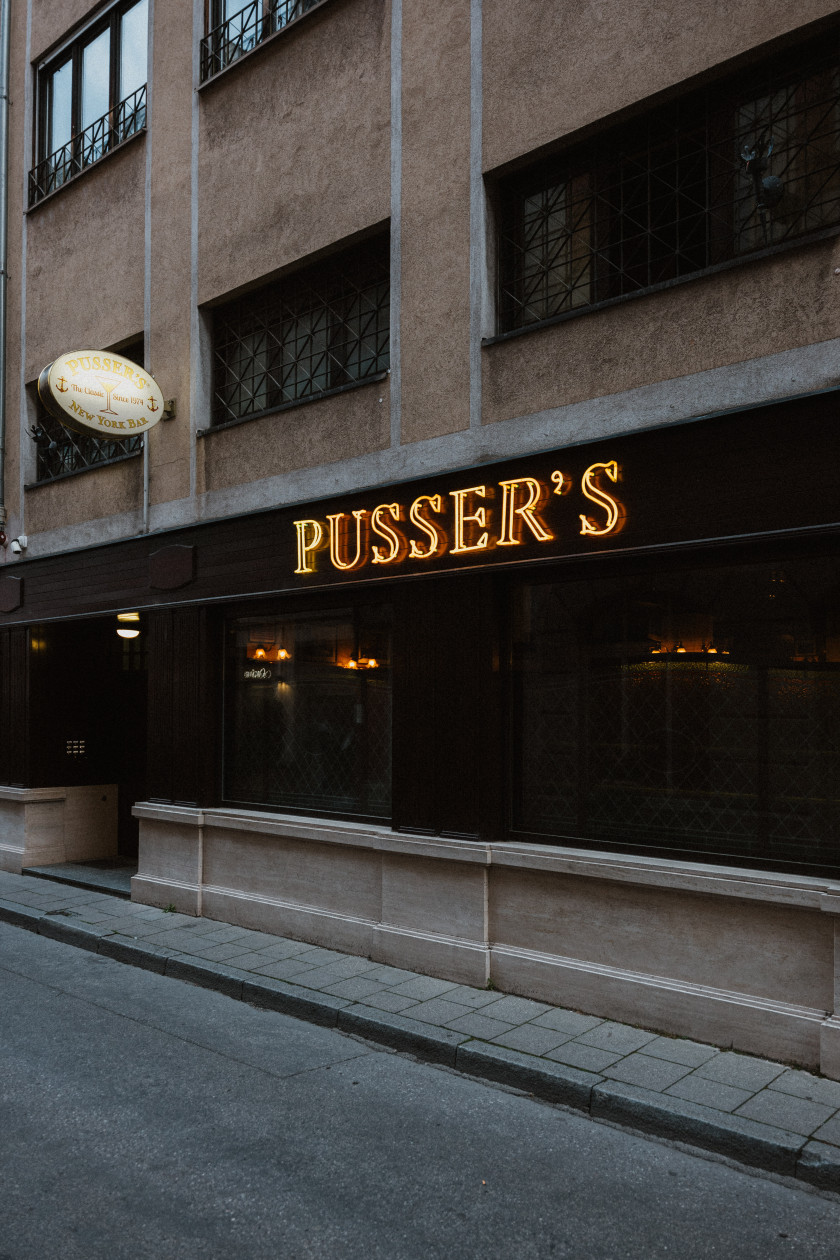 Auf diesem Foto sieht man einen schrägen Blick auf die Außenfassade von der Straße aus. Man erkennt die Straße und den Gehweg vor der Bar. Außerdem sieht man die dunklen Scheiben der Bar. Darüber ist in orangenen Leuchtbuchstaben der Schriftzug „PUSSER´S“ zu lesen.