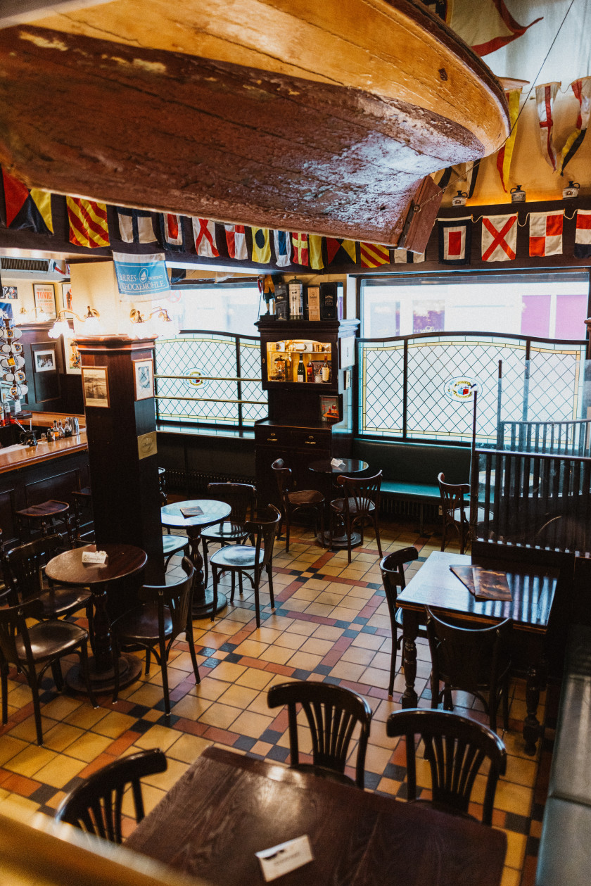 Auf diesem Foto hat man einen Blick in die Bar von einer Erhöhung aus fotografiert. Von der Decke hängt ein Boot aus Holz. Im Innenraum der Bar blickt man auf Tische mit Stühlen oder Bänken. Das Material der Tische und Stühle ist dunkles Holz. Der Boden ist hell gefliest. Im linken Hintergrund des Bildes kann man einen Teil des Tresens erkennen. An den Wänden hängen Girlanden mit verschiedenen Flaggen. Im Hintergrund sind die Fenster der Bar zu erkennen, die fast eine Wandseite einnehmen. Zwischen den beiden großen Fenstern steht ein Regal, in dem und auf dem Flaschen stehen. Vor den Fenstern ist ein Sichtschutz angebracht. Auf den Tischen liegen Karten oder Reservierungsschilder.