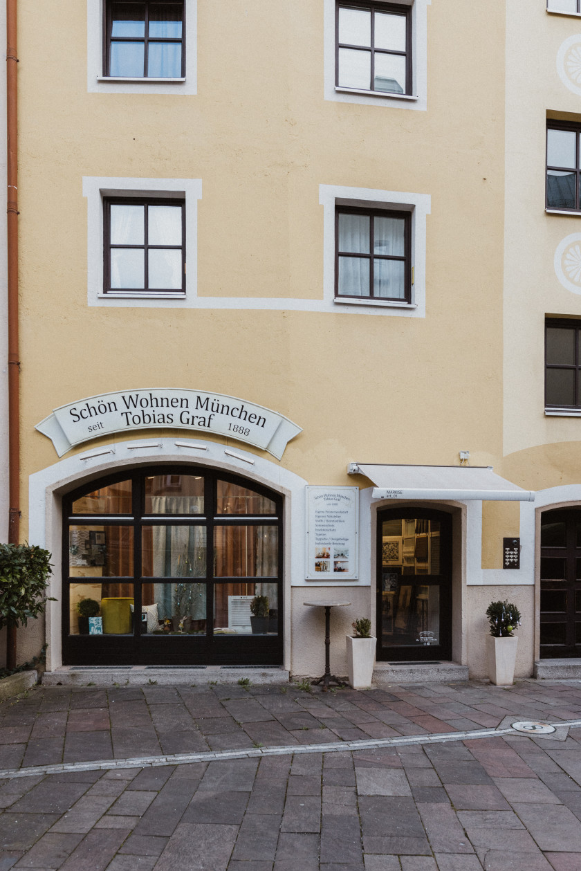 Auf diesem Foto sieht man eine Draufsicht des Ladens von außen aus Perspektive der Asamhöfe. Das Geschäft befindet sich in einem gelben Haus. Rechts im Foto ist die Eingangstür zu erkennen. Rechts und links neben der Einkaufstür befinden sich Blumentöpfe. Zwischen Eingangstür und Schaufenster sind ein Stehtisch und ein Schild mit dem Angebot des Ladens angebracht. Links im Bild ist das Schaufenster zu sehen. Darüber ist ein weißes Schild mit der Aufschrift „Schön Wohnen München Tobias Graf, seit 1888“ angebracht.