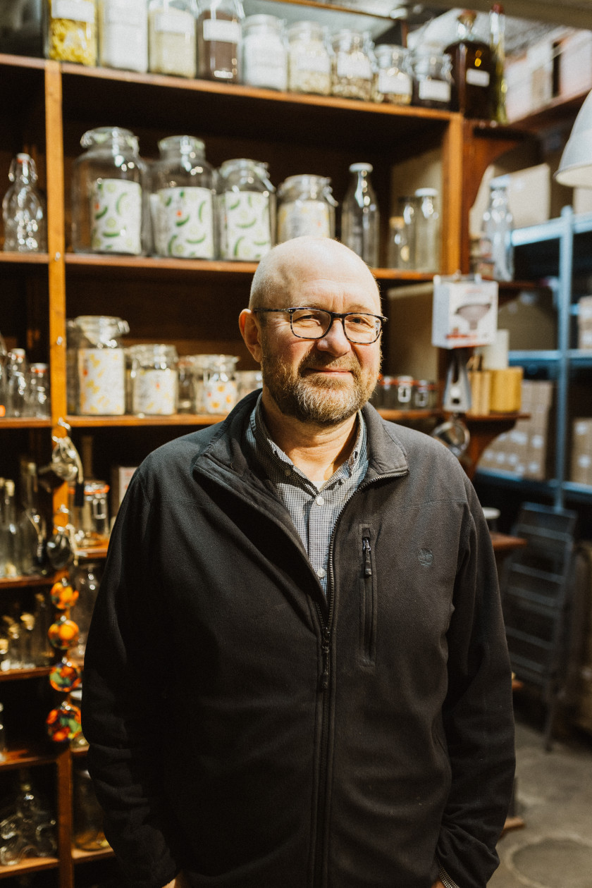 Auf dem Bild ist Peter Ozols zu sehen, der Inhaber des Ladens. Er steht im Laden vor einer Regalwand, gefüllt mit Einmachgläsern, und lächelt in die Kamera. Er hat eine schwarze Jacke über einem blauen Hemd an und trägt eine Brille.