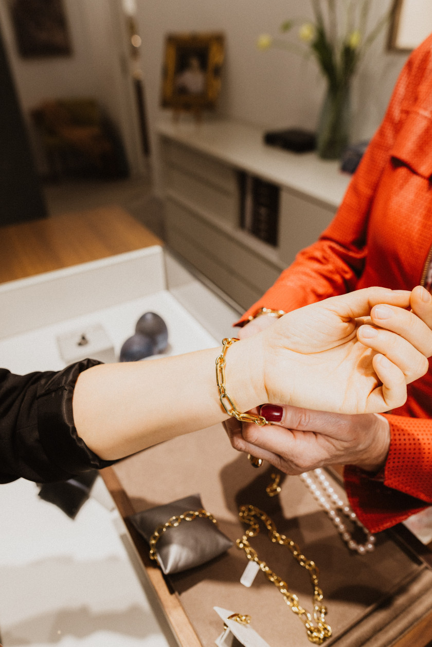 Auf diesem Foto sieht man den Arm einer Frau von links im Bild. Rechts im Bild sieht man einen kleinen Teil eines Oberkörpers, der rot gekleidet ist. Die Hände des Oberkörpers ziehen der Hand, die von links ins Bild ragt, gerade ein goldenes Armband um. Darunter erkennt man Schmuck in einer Vitrine aus Glas liegen.