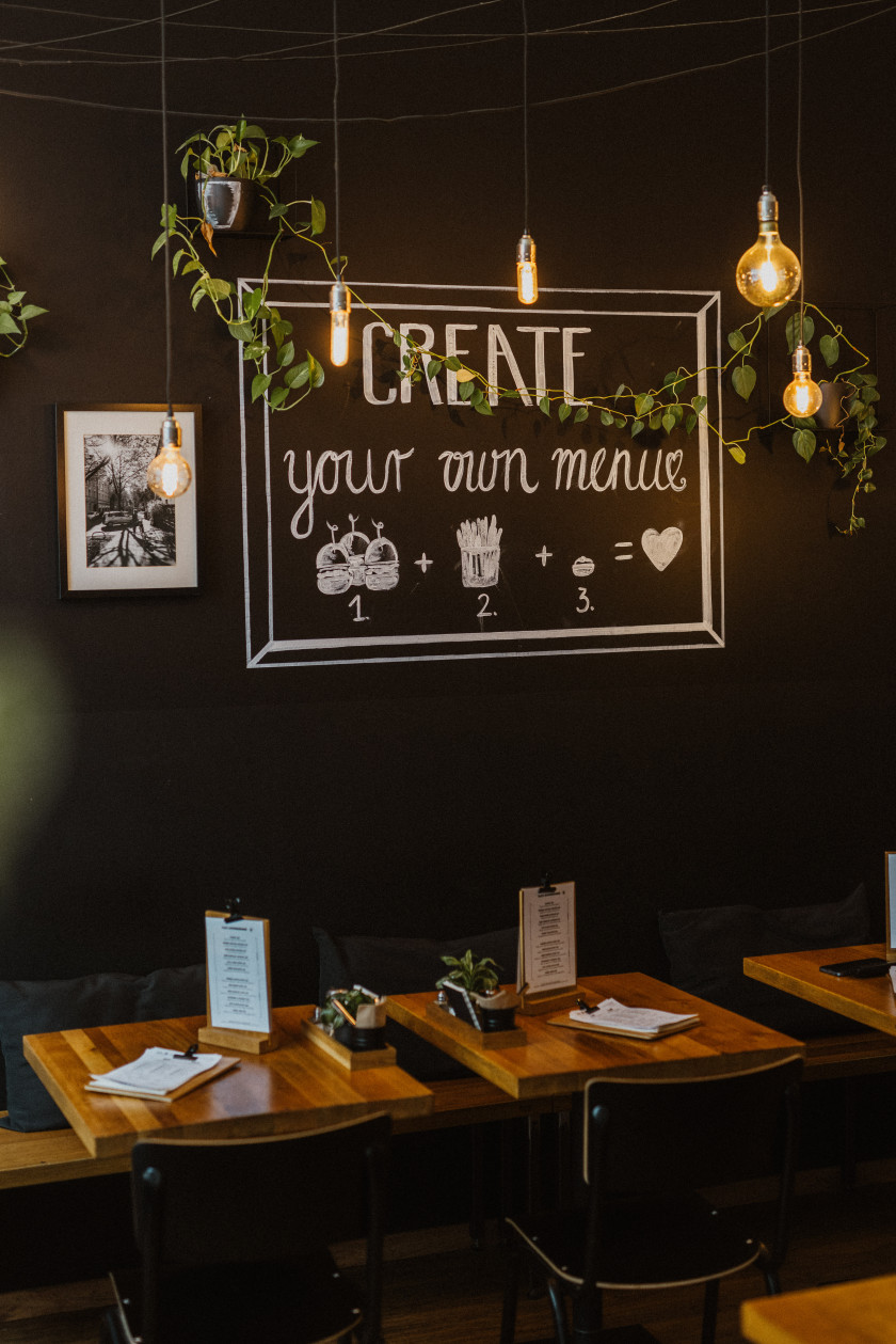 Auf diesem Foto sieht man den Blick auf eine Wand des Restaurants. An der schwarzen Wand steht in weißer Schrift „Create your own menue“. Darunter sind Burger, Pommes und Dips gezeichnet, verbunden mit einem „Plus-Zeichen“. Dahinter ist ein „Gleich-Zeichen“ und dahinter ein Herz. Diese Gleichung könnte man so interpretieren, dass Burger, Pommes und Dips zusammen Liebe ergeben. Daneben hängt ein Foto an der Wand. Von oben hängen Leuchten und Pflanzen in das Foto. Unter dem Bild sind Tische an der Wand zu sehen sowie Bänke an der Wand. Vor den Tischen stehen Stühle. Die Möbel sind in Holzoptik.