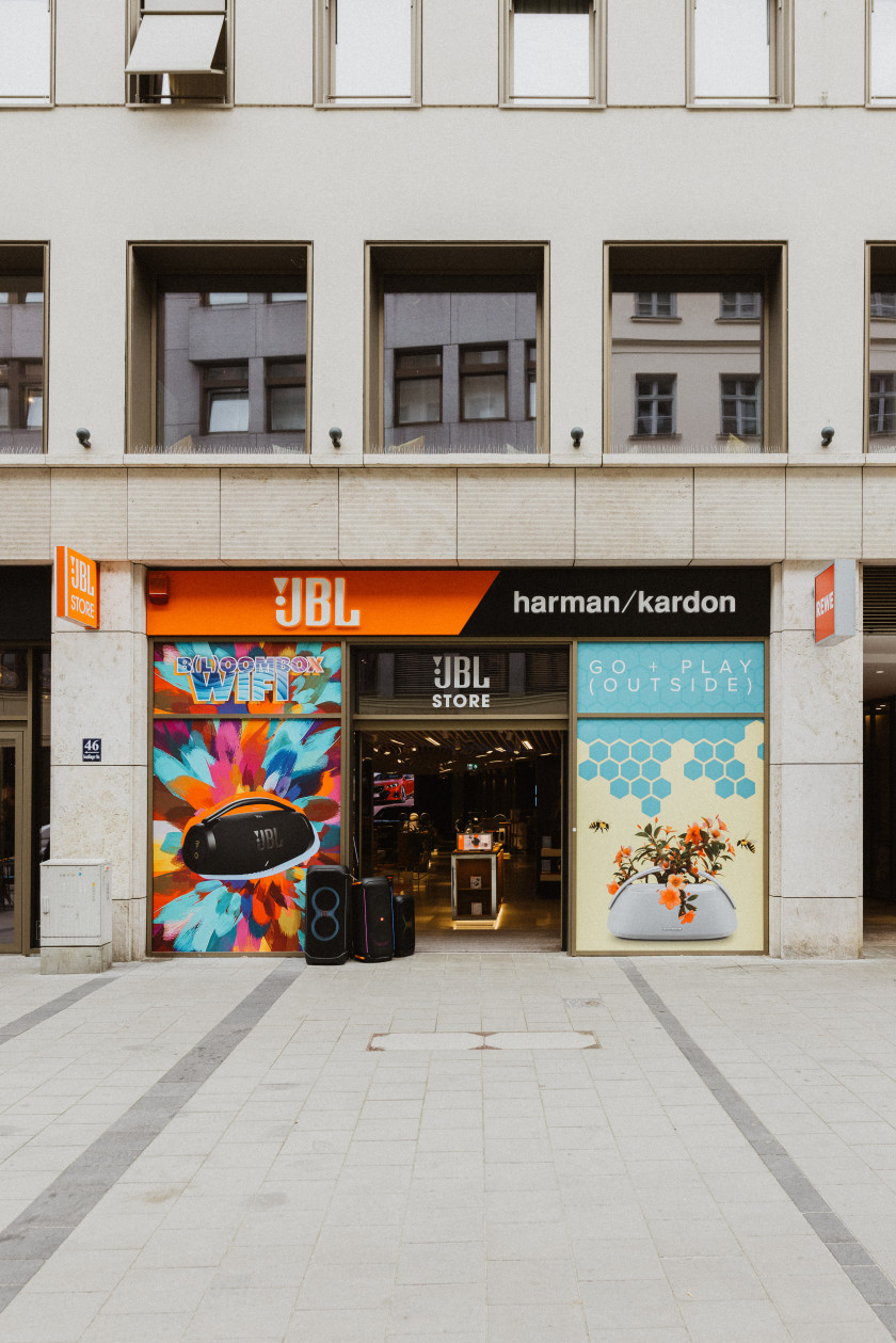 Auf diesem Foto sieht man einen Blick auf die Außenfassade des Geschäfts. Das Haus ist grau. Im Erdgeschoss ist der Eingang zwischen zwei Schaufenstern zu sehen. Die Schaufenster sind beklebt mit Bildern von einem Lautsprecher und einer Blume und Mustern darum. Durch die Eingangstür sieht man einen Blick in den Laden. Vor der Eingangstür stehen Lautsprecher und es ist ein großer Teil der Straße zu sehen. Über der Eingangstür ist „JBL STORE“ zu lesen und darüber „JBL. Harman/kardon“. Darüber sind Fenster im Haus zu sehen.