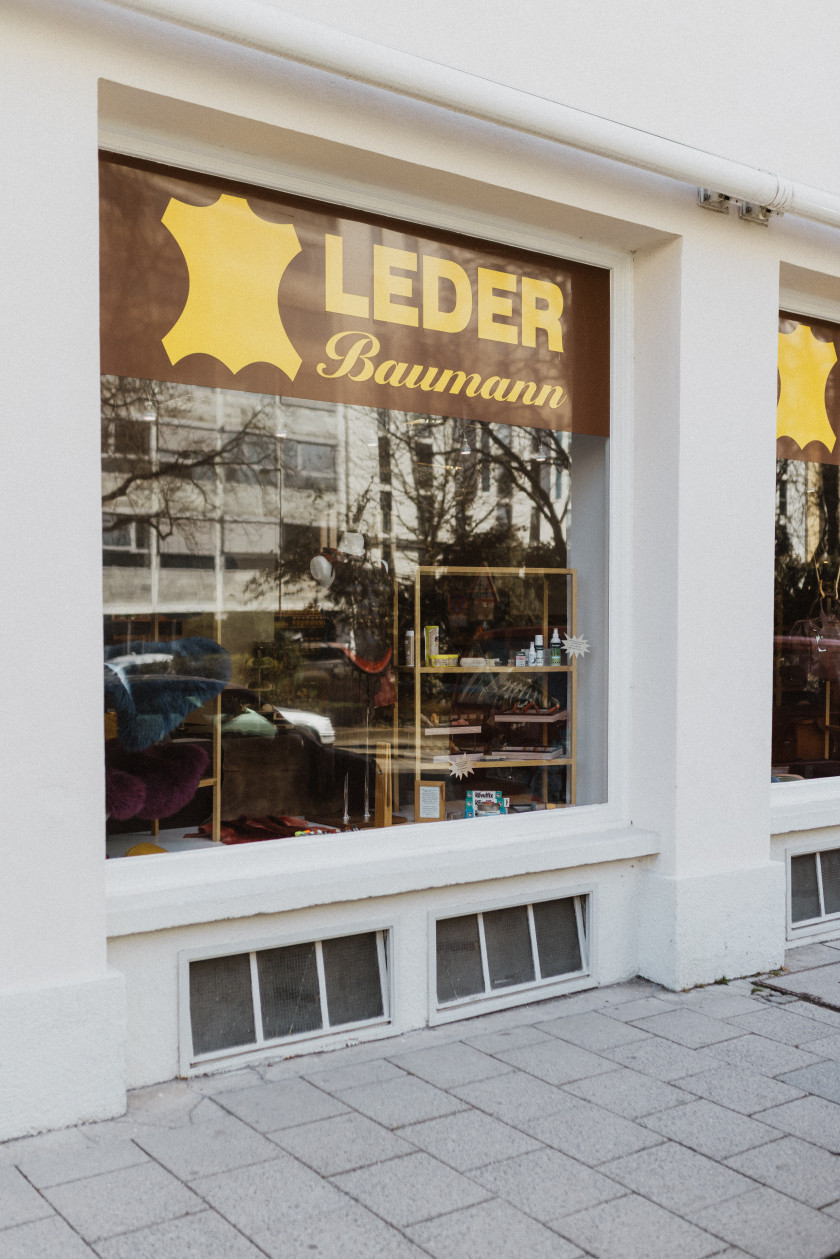 Auf diesem Bild ist die Außenansicht des Geschäftes aus der Perspektive der Herzog-Wilhelm-Straße zu sehen. Man sieht ein Schaufenster und rechts daneben einen kleinen Teil eines weiteren Schaufensters. Auf dem großen Schaufenster steht oben „LEDER Baumann“ in gelber Schrift auf braunem Untergrund. Daneben ist das Logo, das an die Form eines Kuhfells erinnert. Darunter sieht man sehr verspiegelt eine Auslage von Waren im Schaufenster. Unter dem Schaufenster erkennt man zwei Kellerfenster.