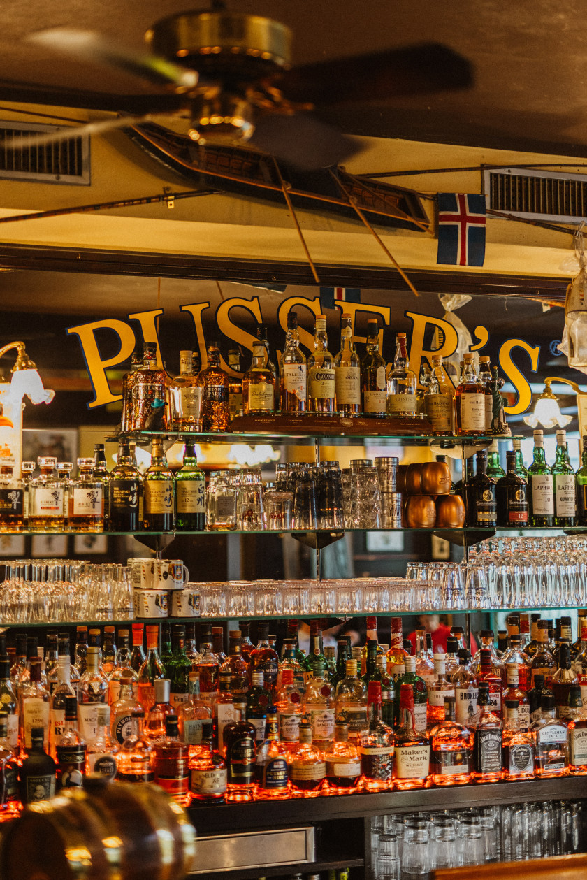 Auf diesem Foto sieht man eine Nahaufnahme der Regale der Bar. Diese sind gefüllt mit verschiedenen Gläsern und Flaschen. Der Teil hinter den gefüllten Regalen ist verspiegelt. Auf dieser Spiegelung ist im oberen Teil der Schriftzug „PUSSER´S“ in Gelb angebracht. Dieser verschwindet zum Teil hinter den Flaschen. Darüber ist noch eine Flagge angebracht. An der Decke hängt ein Ventilator.