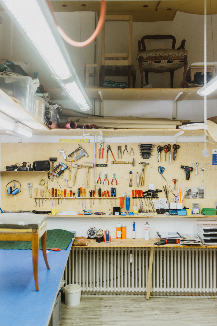 Auf diesem Foto sieht man eine gut organisierte Werkstatt. An der Wand hängen zahlreiche Werkzeuge ordentlich sortiert, darunter Hammer, Schraubenzieher, Zangen, Sägen und andere Handwerksutensilien. Auf der linken Seite des Bildes ist ein Arbeitstisch zu sehen, auf dem ein Stuhl oder ein Hocker auf einer blauen Unterlage steht. Im oberen Bereich des Bildes befinden sich eine Ablagefläche oder ein Hochregal, auf dem weitere Möbelstücke und Materialien lagern, darunter weitere Stühle. Die Werkstatt ist gut beleuchtet, mit mehreren Leuchtstofflampen, die den Arbeitsbereich erhellen. Der Boden besteht aus hellem Holz und unter dem Arbeitstisch ist eine Heizungsanlage zu sehen.