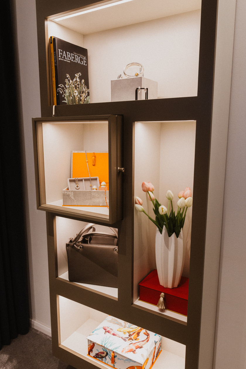 Auf diesem Foto sieht man den Blick in eine Vitrine mit Glasscheiben. In der Vitrine stehen Blumen, Taschen, Schmuckstücke, Bücher und Schachteln, die beleuchtet werden.