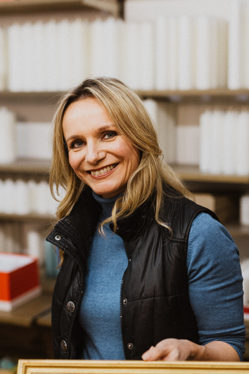 Auf diesem Foto sieht man ein Porträt der Inhaberin Claudia Slanzi. Sie trägt einen blauen Pullover und darüber eine schwarze Weste. Man sieht im untersten Teil des Bildes, dass sie ein Bild in der Hand hält, sieht allerdings nur leicht den Holzrahmen des Bildes. Sie lächelt in die Kamera. Im Hintergrund sind verschwommen Kerzenrohlinge zu sehen.