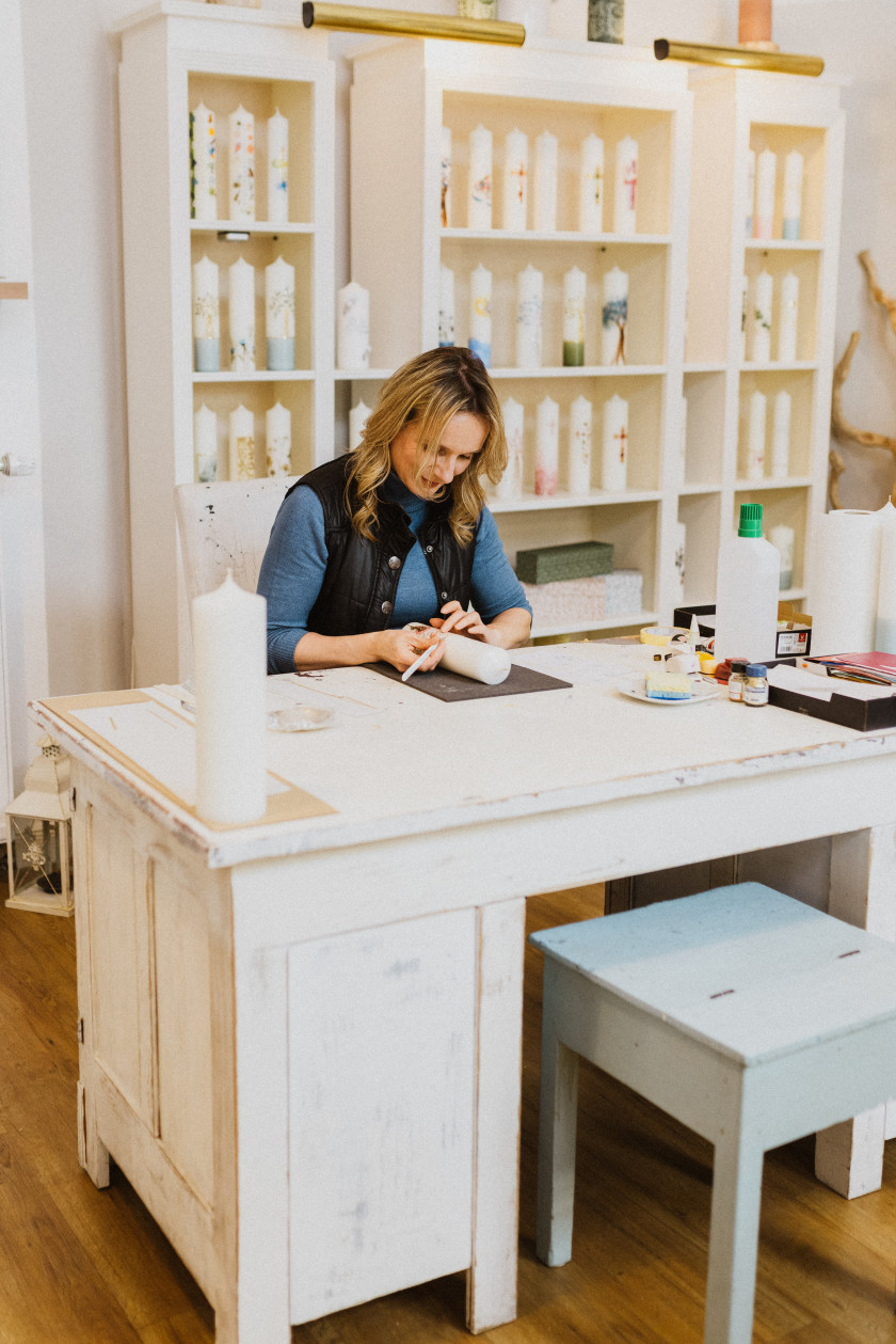 Auf diesem Foto sieht man die Inhaberin Claudia Slanzi, wie sie an einem Werktisch im Laden sitzt. Im Hintergrund ist ein Regal voller verschiedener Kerzen mit unterschiedlichen Designs. Sie sitzt hinter dem Werktisch. Vor dem Werktisch steht ein kleiner Hocker. Der Werktisch ist weiß und aus Holz. Links im Bild auf dem Werktisch steht eine große weiße Kerze. Claudia Slanzi ist vertieft in ihre Arbeit und bearbeitet gerade mit beiden Händen eine Kerze. Rechts im Bild auf dem Werktisch stehen weitere Utensilien zur Bearbeitung von Kerzen.