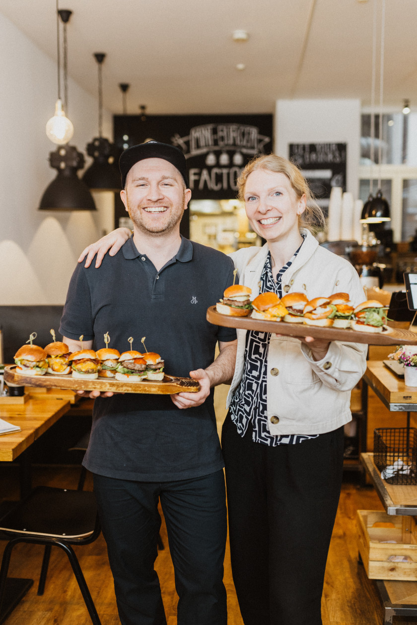 Auf diesem Foto sind die beiden Inhaber zu sehen. Links im Bild steht Florian Kantor und rechts im Bild daneben steht Julia Kantor. Die beiden haben Holztabletts mit Miniburgern in den Händen. Julia Kantor hat einen Arm um Florian Kantor gelegt und in dem anderen Arm hält sie das Tablet mit den Burgern. Die beiden lächeln in die Kamera. Florian Kantor trägt eine schwarze Cap, ein dunkelgraues Polo-Shirt und schwarze Hosen. Julia Kantor trägt eine gemusterte Bluse, darüber eine weiße Jacke und dazu schwarze Hosen. Ihre Haare sind zusammengebunden. Im Hintergrund ist das Restaurant verschwommen zu erkennen. Das Foto wurde in Richtung der Küche aufgenommen.
