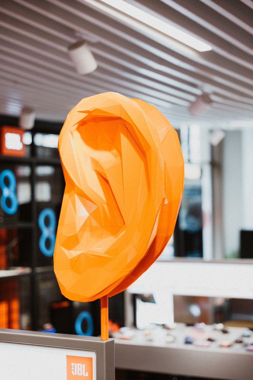 Auf dem Foto ist ein Ohr aus Plastik oder Kunststoff in Nahaufnahme zu sehen. Das Ohr ist orange und an einem Aufsteller befestigt. Im Hintergrund ist verschwommen ein Teil des Ladens zu sehen.