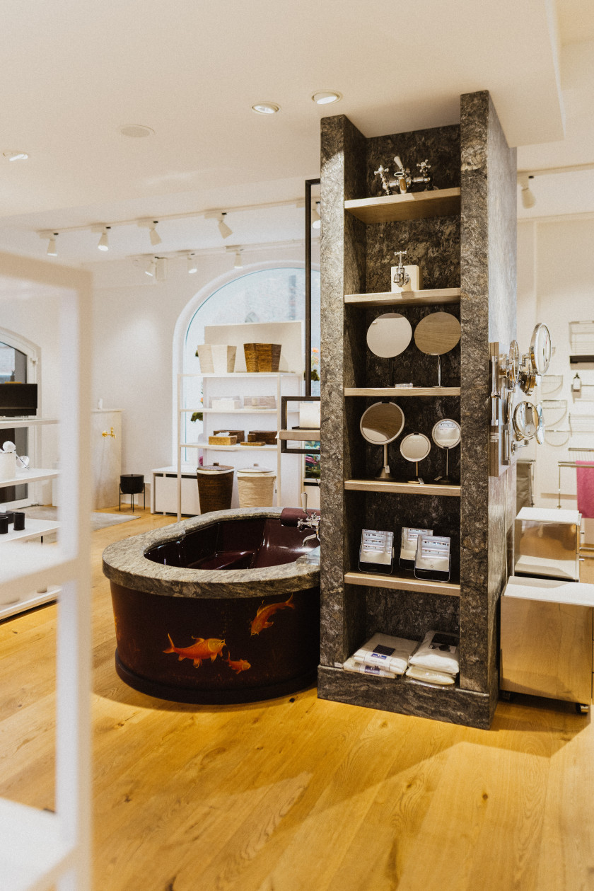 Auf diesem Foto sieht man eine Badewanne links neben einem deckenhohen Regal aus Marmor stehen. Die Badewanne ist schwarz und darauf sind Goldfische zu sehen. In dem deckenhohen Regal aus Marmor rechts daneben stehen verschiedene Bad-Accessoires wie Spiegel oder Armaturen für Wasserhähne. Seitlich an diesem Regal ist zu erkennen, dass ebenfalls Spiegel daran befestigt sind. Im Hintergrund ist der Verkaufsraum zu erkennen.