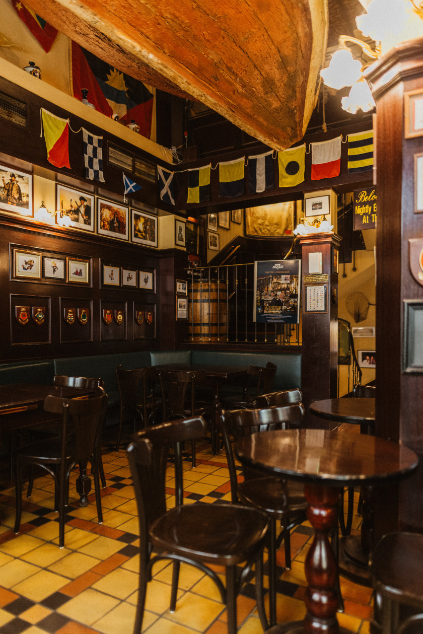 Auf diesem Foto sieht man einen Blick in die Bar. Von der Decke hängt ein Boot aus hellem Holz. Oben an den Wänden sind außerdem Girlanden mit verschiedenen Flaggen befestigt. Links an den Wänden sind verschiedene Bilder aufgehängt. Im Hintergrund ist eine Treppe, die nach unten und nach oben führt, zu erkennen. Im Vordergrund sind die Tische mit Stühlen aus dunklem Holz. Der Boden ist hell gefliest. Auch an den beiden erkennbaren Holzbalken im Raum sind Bilder und Lampen befestigt.