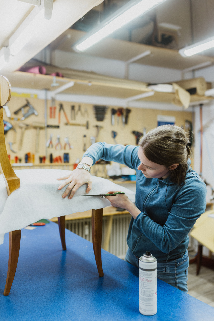 Auf diesem Foto sieht man eine junge Frau, die an einem Stuhl arbeitet. Der Stuhl steht auf einer blauen Arbeitsplatte links im Foto. Der Stuhl ist aus Holz und die Sitzfläche ist mit einem weißen Stoff bedeckt. Die junge Frau steht vor der Arbeitsplatte mit dem Stuhl und hält eine Schere in der einen Hand, mit der sie gerade den weißen Stoff auf der Sitzfläche des Stuhls bearbeitet. Mit der anderen Hand hält sie den weißen Stoff fest. Es sieht so aus, als würde der Stuhl gerade gepolstert werden. Auf der Arbeitsfläche steht außerdem eine Sprühflasche. Die junge Frau blickt konzentriert auf den Stoff. Sie trägt ein blaues Sweatshirt. Im Hintergrund sind an der Wand aufgehängt verschiedene Werkzeuge zu erkennen, wie Zangen oder Hammer. Darunter ist ein Heizkörper zu sehen.