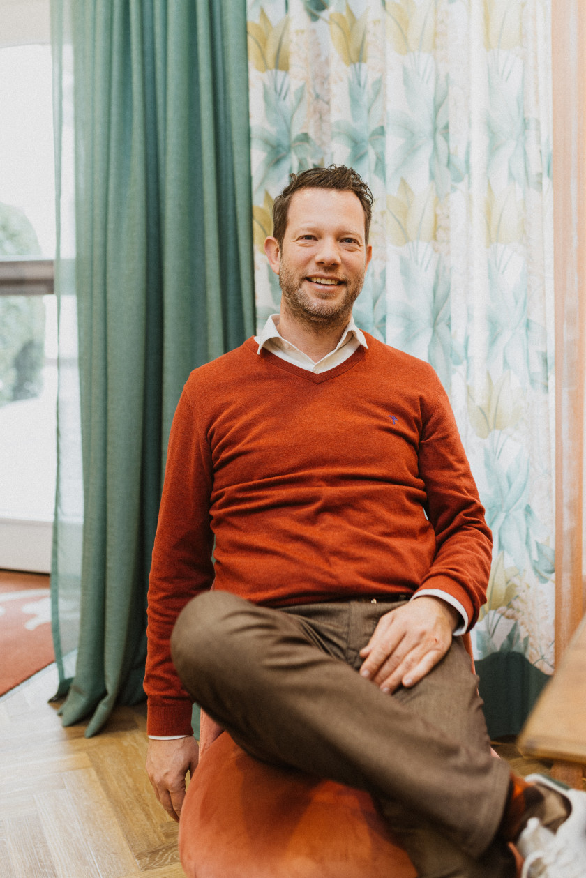 Auf diesem Foto ist der Inhaber Tobias Graf zu sehen. Er sitzt auf einem orangenen Sessel im Laden. Hinter ihm sind verschiedene Vorhänge aufgehängt zu sehen. Er hat ein Bein über das Andere geschlagen. Er lächelt in die Kamera und trägt ein rotes Sweatshirt. Darunter schaut der Hemdkragen hervor. Dazu trägt er eine braune Stoffhose, weiße Sneaker und braun-rot gemusterte Socken. Eine Hand hat er auf seinem Oberschenkel und der andere Arm hängt neben seinem Körper.