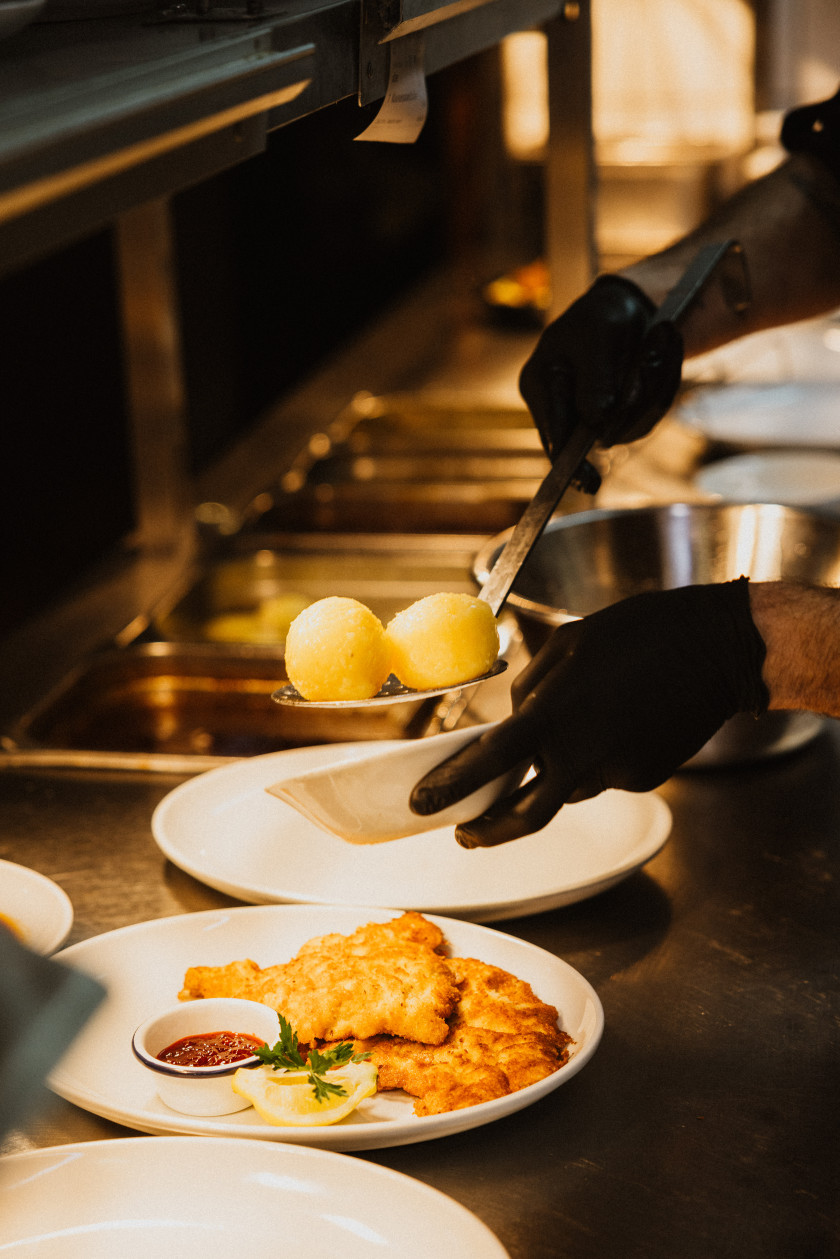 Auf diesem Foto ist eine Aufnahme in der Küche zu sehen. Man sieht die silberne Küchentheke. Auf dieser stehen vier Teller. Zwei Teller sind ganz zu erkennen. Auf dem einen Teller liegen zwei Schnitzel mit einer Zitrone und Preiselbeeren in einem Schüsselchen. Der andere Teller ist noch leer. Über dem Teller sieht man zwei Hände in schwarzen Handschuhen. In der einen Hand hält die Person zwei Knödel auf einem Schöpfgerät. In der anderen Hand hält die Person eine Schüssel. Die Knödel werden gerade in die Schüssel gelegt. Im Hintergrund sieht man noch eine silberne Schüssel und Aufbewahrungsbehälter. Über der Theke ist eine schwarze Schiene, an der ein weißes Zettelchen hängt.