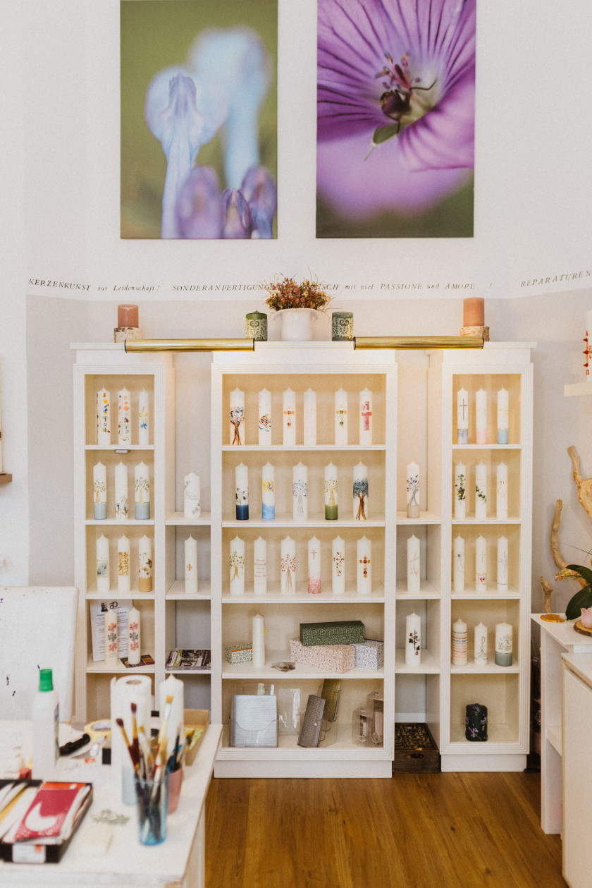 Auf diesem Foto ist der Blick in den Laden auf eine Wand mit einer Regalfront zu sehen. Ganz oben an der Wand sind zwei Fotografien von lilafarbenen Blüten. Darunter steht eine große Vitrine. Auf und in der Vitrine stehen handverzierte Kerzen. Alle in einer Größe, aber mit unterschiedlichen Verzierungen. Unten in der Vitrine stehen auch Schachteln bzw. Boxen und andere Dinge. Links vorne im Vordergrund des Fotos sieht man eine Ecke des weißen Werktisches, auf dem Pinsel, Zewarolle und weitere Utensilien zur Bearbeitung von Kerzen stehen.