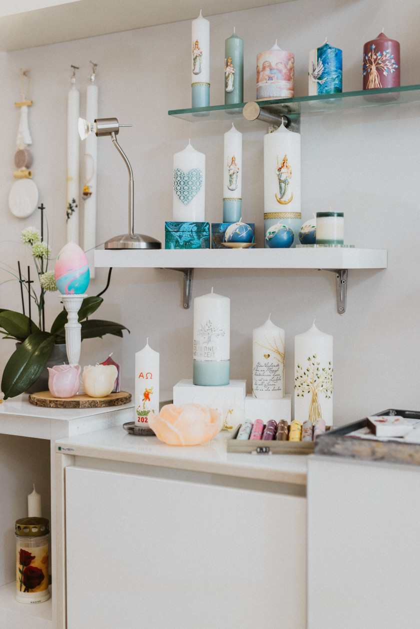 Auf diesem Foto sieht man den Ausschnitt einer Wand des Ladens. An der Wand hängen Regale und unter den Regalen ist ein Sideboard zu sehen. Das oberste Regal ist aus Glas und darauf sind verschieden verzierte Kerzen zu sehen. Die Kerzen sind bunt und haben unterschiedliche Formen. Das Regal darunter ist aus weißem Holz und darauf befinden sich ebenfalls Kerzen in unterschiedlichen Farben, Formen und Designs. Links im Bild auf diesem Regal steht eine Lampe und links neben dem Regal hängen Kerzen an der Wand. Auf dem Sideboard stehen und liegen weitere Kerzen und links auf dem Sideboard eine Blume. Auch besondere Kerzen in Form von Blüten sind auf dem Regal zu erkennen.
