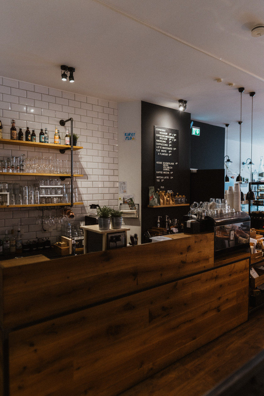 Auf diesem Foto ist die Theke des Restaurants zu sehen. Die Theke ist aus braunem Holz. Dahinter kann man ein Regal an einer Wand mit weißen Fliesen erkennen. An der Decke sind Strahler angebracht. In dem Regal stehen verschiedene Gläser und Flaschen. Rechts an der Theke im Bild ist eine Kaffeemaschine zu erkennen, auf der ebenfalls Gläser und To-go-Becher stehen. Rechts im Hintergrund an der schwarzen Wand, die sich rechts im Bild neben der weiß gefliesten Wand befindet, ist das Menü mit weißer Kreide auf eine schwarze Tafel geschrieben. Daneben ist ein Exit-Schild an der Decke.