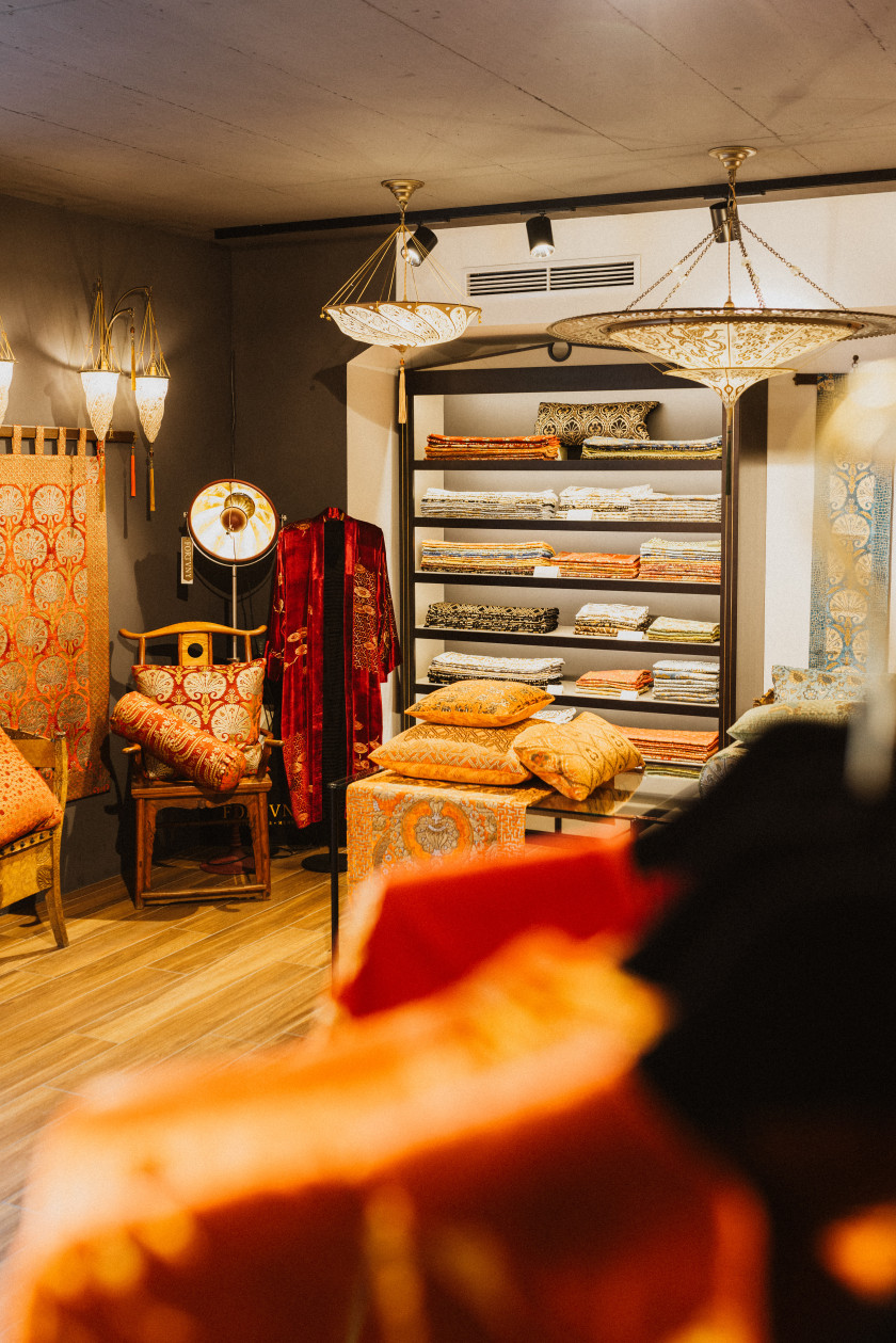 Dieses Bild wirft einen Blick in den Laden. Im Vordergrund sind rechts verschwommene Kleidungsstücke in Orange. Im Hintergrund sind ein Regal mit Stoffen, ein Stuhl mit Kissen, Strahler, ein Wandteppich und Leuchten an der Decke zu sehen.