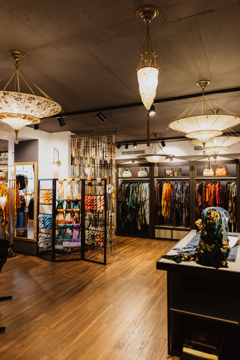 Auf diesem Foto sieht man den Blick in den Verkaufsraum. An der Decke hängen die Deckenleuchten. Im Hintergrund hängen die Kimonos an den Kleiderbügeln, darüber stehen Taschen im Regal. Zudem sind viele weitere Details im Hintergrund zu finden: ein Tisch mit Tüchern, ein Spiegel, ein Aufsteller mit den Seidensträngen.