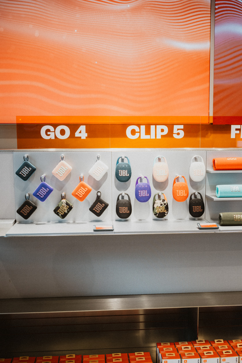 Auf diesem Foto sieht man eine Wand des Geschäfts, an der Lausprecher in verschiedenen Farben und Formen befestigt sind. Über den Lautsprecher steht „GO 4, CLIP 5“ in weißem Schriftzug. Darüber ist eine orange Wand zu sehen.
