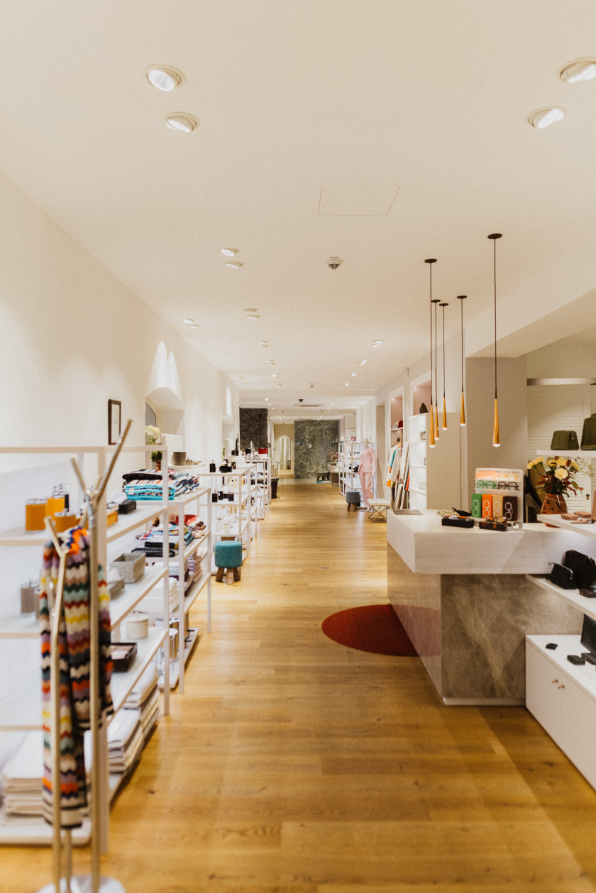 Auf diesem Foto sieht man einen tiefen Blick in den Laden. Rechts seitlich im Bild ist der Verkaufstresen zu erkennen. Links seitlich im Bild sind weiße Regale mit verschiedenen Bad-Accessoires wie Seifenspendern oder Handtüchern zu erkennen. Dazwischen führt ein breiter und sehr langer Gang in den hinteren Teil des Ladens.