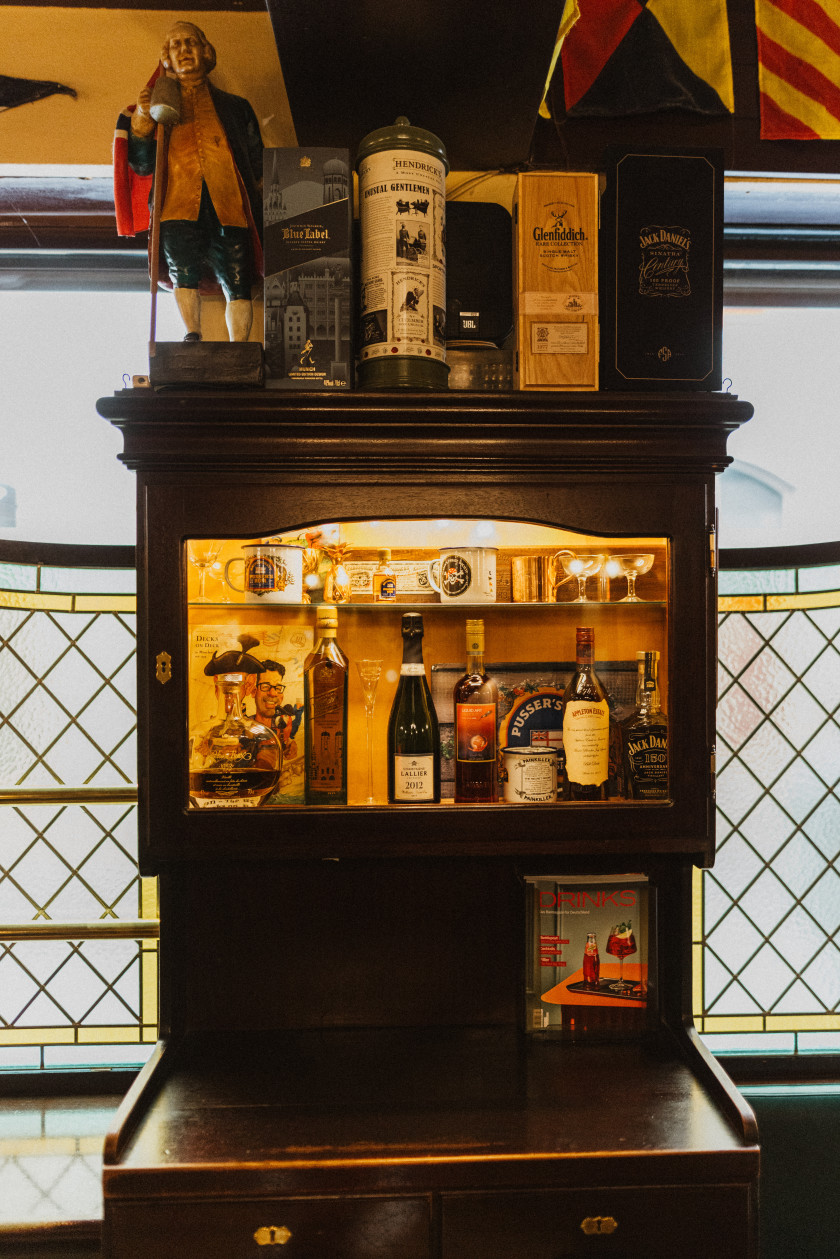 Auf diesem Foto steht eine Vitrine aus dunklem Holz im Vordergrund. Links und rechts von der Vitrine sind Fenster erkennbar, durch die das Tageslicht scheint. Unten auf der Vitrine steht ein Buch. Darüber stehen hinter Glas verschiedene Flaschen, Bücher und Trinkgefäße, die angestrahlt werden. Darüber stehen ebenfalls Flaschen in ihren Verpackungen und die Figur eines Mannes.