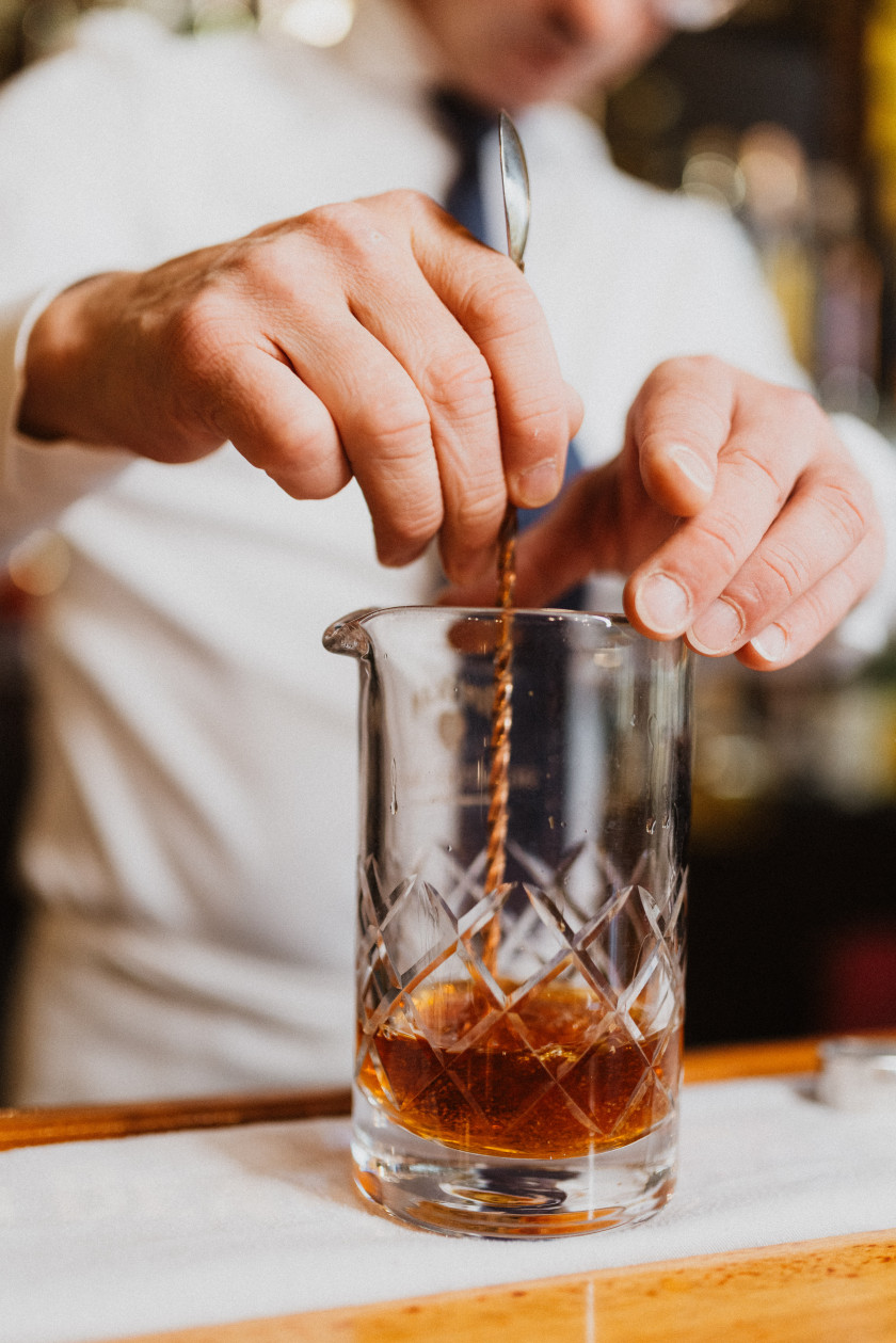 Auf diesem Foto sieht man ein durchsichtiges Glas im Fokus. In dem Glas befindet sich eine braune Flüssigkeit und das Glas ist rund ein Viertel gefüllt. Man kann den Oberkörper von David Deck im Hintergrund durch das weiße Hemd und die schwarze Krawatte verschwommen erkennen. Den Kopf sieht man nicht. Mit der einen Hand hält er das Glas fest und in der anderen Hand hält er den Barlöffel und rührt damit die Flüssigkeit im Glas um. Das Glas steht auf einer weißen Serviette auf dem Tresen aus hellem Holz.