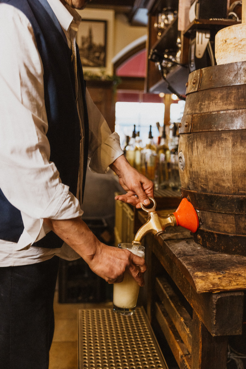 Auf diesem Foto sieht man, wie ein Mann, dessen Kopf man nicht sieht, vor einem Bierfass steht. Der Mann trägt ein weißes Hemd mit Trachtenweste. Mit der einen Hand dreht er am Zapfhahn, der im Fass steckt, und in der anderen Hand hält er ein Bierglas unter den Zapfhahn. Das Bier fließt aus dem Zapfhahn in das Glas. Das Glas ist bereits fast voll mit Schaum. Im Hintergrund ist der Thekenbereich zu erkennen.