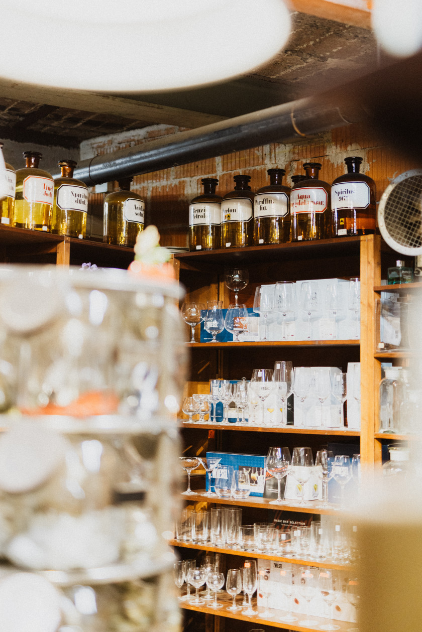 Auf diesem Bild sieht man ein Regal mit verschiedenen Glasarten gefüllt. Ganz oben drauf stehen große Apothekerflaschen aus braunem Holz. Darunter verschiedene Trink- und Weingläser sortiert nebeneinander. Links im Vordergrund ist verschwommen ein Aufsteller zu erkennen. Im Hintergrund sind oben an der Decke eine unverputzte Wand und ein Stahlrohr zu erkennen.