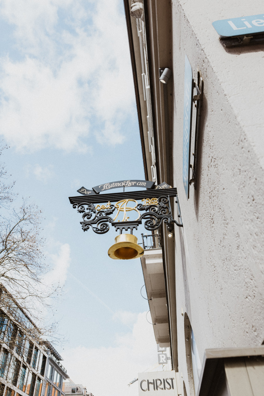 Auf diesem Foto sieht man die Außenansicht von Hut-Breiter. Im Fokus steht in Nahaufnahme aufgenommen ein Schild, das vor dem Laden über den Schaufenstern befestigt ist. Das Schild steht raus und senkrecht zur Außenfassade. Auf dem Zeichen ist ein goldener Hut in Miniaturformat befestigt und darüber steht in goldener Schrift auf schwarzem Hintergrund „Der Hutmacher am Dom“. Rechts im Bild sieht man die Außenfassade seitlich. Links im Bild sieht man den Teil eines Gebäudes seitlich. Mittig im Bild sieht man den blauen Himmel mit Wolken, da das Bild von unten aufgenommen wurde.