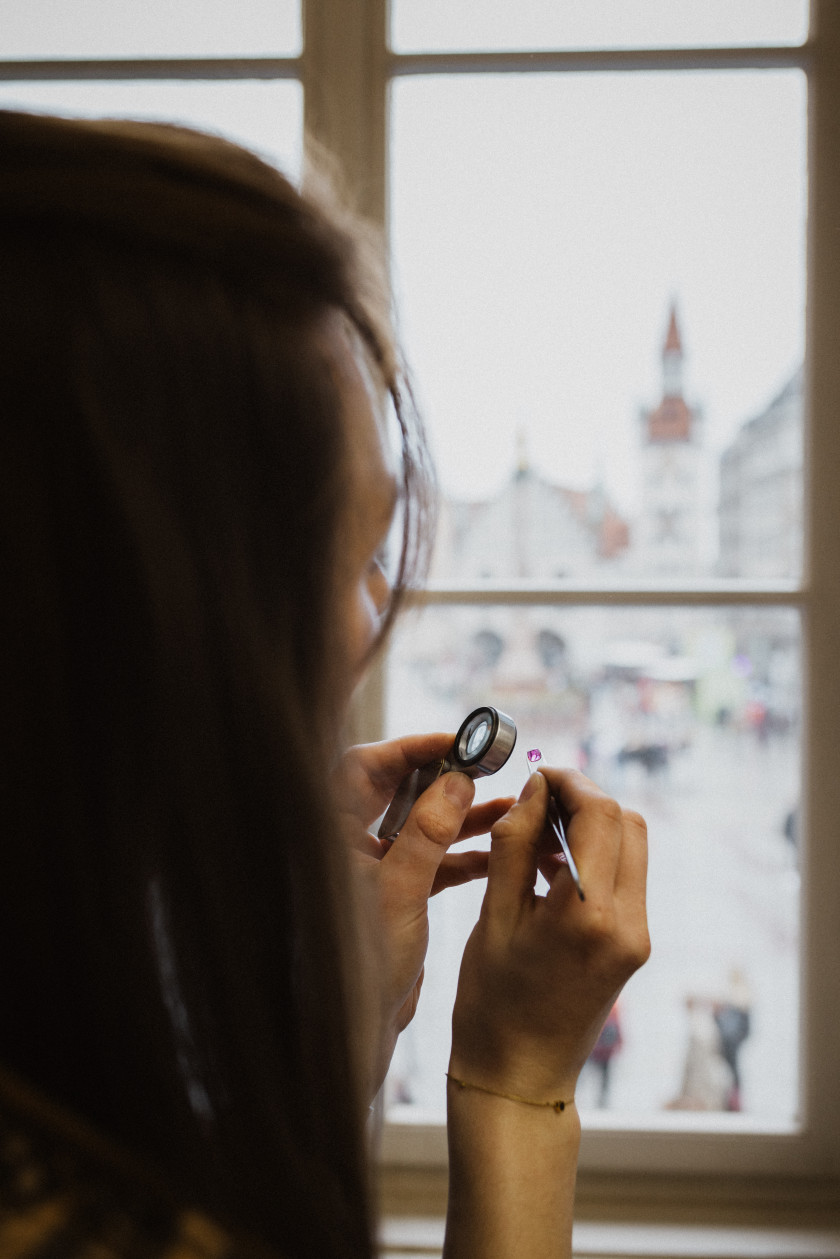 Auf diesem Foto wird der Kopf einer Frau von hinten fotografiert, sodass man nur ihre Haare sieht. Vor sich auf Kopfhöhe hält sie in der einen Hand eine Lupe und in der anderen Hand eine Pinzette mit einem Stein. Im Hintergrund ist durch ein Fenster ein Teil des Stadtbildes zu erkennen.
