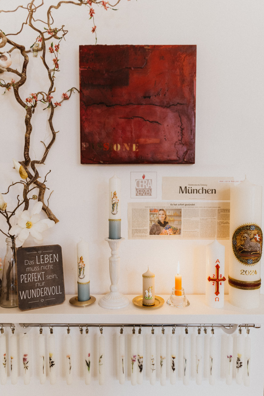 Auf diesem Foto ist ein Teil der Wand des Ladens zu sehen. Oben an der Wand im Bild hängt ein rotes Gemälde. Links daneben im Bild steht ein Ast mit Blüte und darunter das Schild „Das Leben muss nicht perfekt sein, nur wundervoll.“ Links daneben stehen Kerzen auf Kerzenständern auf einem Regal. Eine orangene Kerze brennt in der Mitte. Auch rechts im Bild stehen zwei Kerzen auf dem Regal. Unter dem Gemälde und über den Kerzen ist ein Zeitungsartikel aus der Süddeutschen Zeitung aufgehängt, auf dem die Inhaberin Claudia Slanzi und ein Text zu sehen sind. Daneben hängt noch in etwas kleiner ein Sticker mit der Aufschrift „CERA KERZEN“ untereinander, sodass es aussieht wie eine Kerze, und oben drüber ist noch eine Flamme. Unter dem Regal ist eine Schiene angebracht, an der viele Blumenkerzen aufgehängt sind.