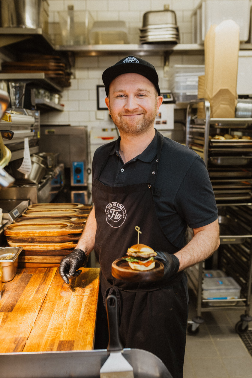 Auf diesem Foto sieht man Florian Kantor im Zentrum des Bildes. Er trägt eine schwarze Cap, ein schwarzes Polo-Shirt und darüber eine schwarze Schürze. Er lächelt. In seiner linken Hand hält er einen Mini-Burger auf einem kleinen Tablett aus Holz. Er steht vor einer Küchentheke aus Holz. Im Hintergrund ist eine Küche zu erkennen.