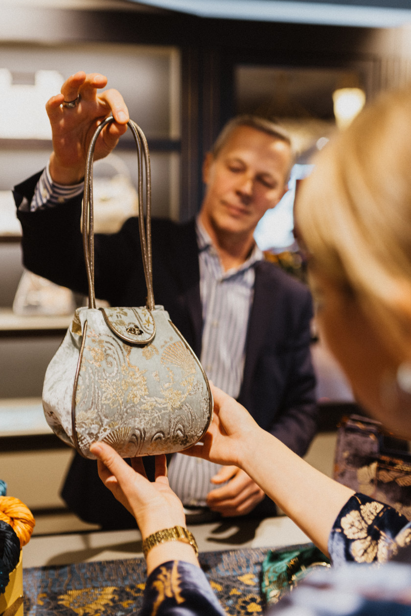 Ein Verkäufer zeigt einer Kundin eine exklusive Tasche. Das Foto ist durch einen Schulterblick aufgenommen. Im Hintergrund sieht man den Verkäufer verschwommen, wie er die Tasche hochhält, und diese ist im Zentrum des Bildes. Die Tasche ist blau gemustert. Die Kundin nimmt die Tasche leicht in die Hand.