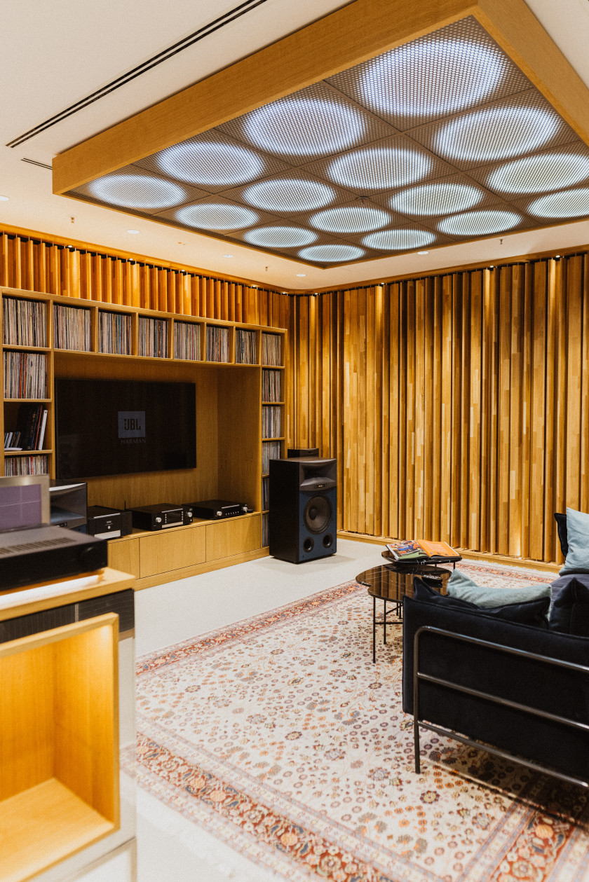 Auf diesem Foto ist ein Blick in einen Raum, der eingerichtet ist wie ein Wohnzimmer, zu sehen. Die Wände sind mit Holz verkleidet. An einer Wand ist ein TV zu sehen und um den TV herum ein Regal mit Platten. Davor steht ein großer Lautsprecher. Am Boden liegt ein Teppich. Auf dem Teppich sieht man einen kleinen Tisch und dahinter ist ein Teil einer Couch zu sehen.