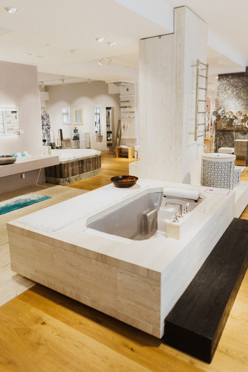 Auf diesem Foto ist eine Badewanne mit breiter integrierter Sitz- oder Liegefläche aus Marmor im Mittelpunkt. Die Badewanne steht im Verkaufsraum. Dahinter sind weitere Verkaufsprodukte im Laden zu erkennen wie Badewannen, Armaturen etc. Der Raum ist sehr hell und gut beleuchtet.