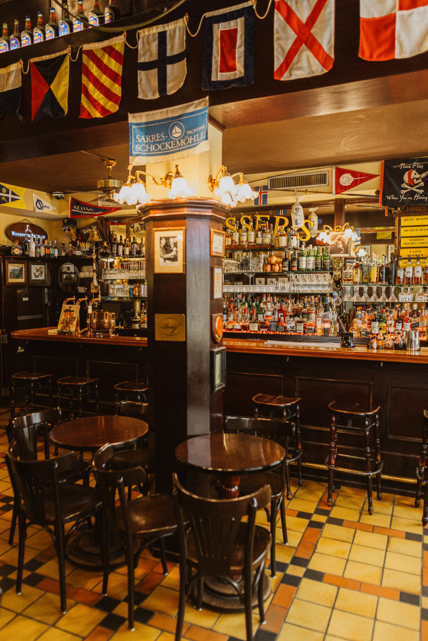 Auf diesem Foto sieht man einen Blick in den Innenraum der Bar und auf die Bartheke. Im Vordergrund sind Stühle und runde Tische aus dunklem Holz, die vor einem Holzbalken, der mitten im Raum steht, stehen. An dem Holzbalken sind Bilder und eine Lampe befestigt. Der Boden ist hell gefliest. Hinter dem Holzbalken ist der Tresen zu sehen. Vor dem Tresen stehen Barhocker, ebenfalls aus dunklem Holz. Dahinter ist die Bar gefüllt mit verschiedenen Flaschen und Gläsern zu erkennen. Über dem Balken an der Wand hängt eine Girlande mit verschiedenen Flaggen.