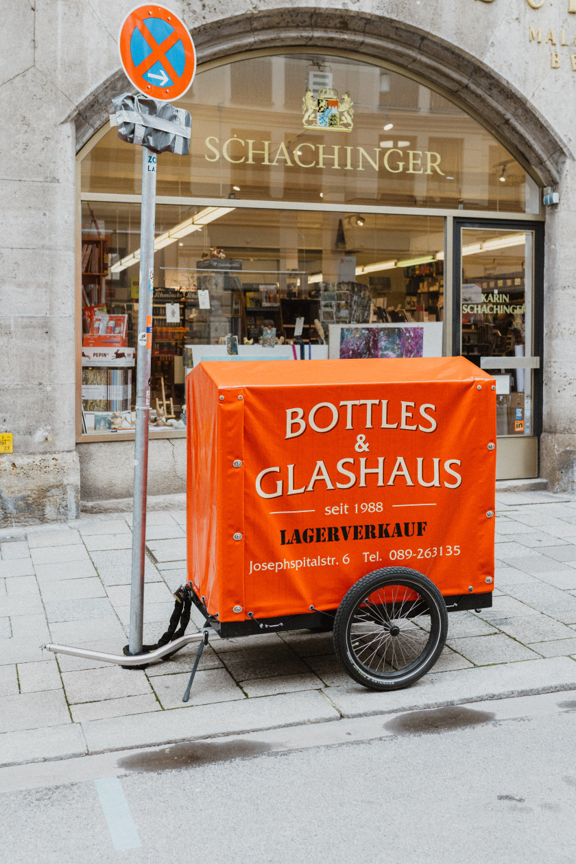Auf diesem Foto sieht man einen Fahrradanhänger. Auf dem Fahrradanhänger ist eine rote Plane, auf der Folgendes steht: Bottles & Glashaus, seit 1988, Lagerverkauf, Josephspitalstr. 6, Tel.: 089-263135. Man sieht ein Rad und es ist eine Draufsicht von der Seite. Im Hintergrund sieht man das Schaufenster von Schachinger. Der Anhänger steht auf dem Gehweg vor dem Hinterhof.