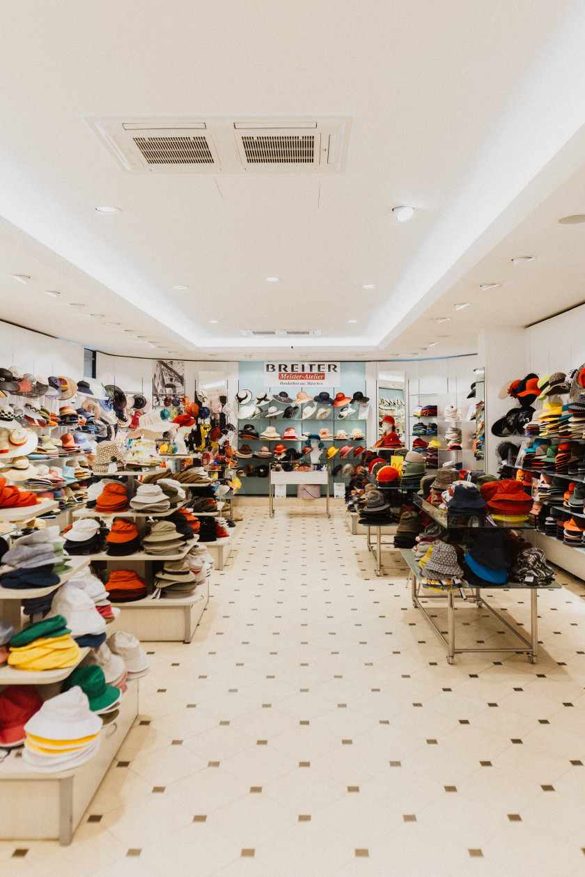 Auf diesem Foto sieht man einen Blick in das Geschäft, konkreter in den 1. Stock des Ladens. Hier sind viele Frauenhüte auf Auslagentischen oder dahinter in Regalen zu sehen. Die Frauenhüte haben alle verschiedene Formen und Farben. Im Hintergrund ist in der Ferne das Ende des Verkaufsraums zu sehen. Die Wand ist türkis und ganz oben steht der Schriftzug „Breiter, Meisteratelier“. Der Boden ist weiß gefliest mit schwarzen kleinen Quadraten.